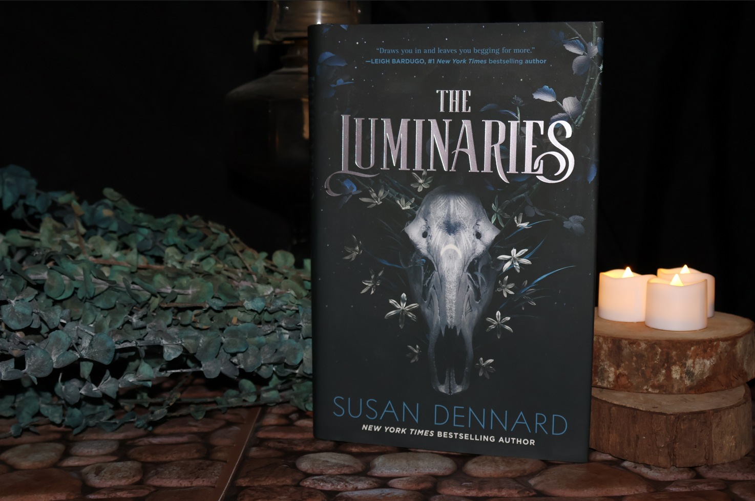 The Luminaries book, framed by candles, greenery, and on a cobblestone surface.