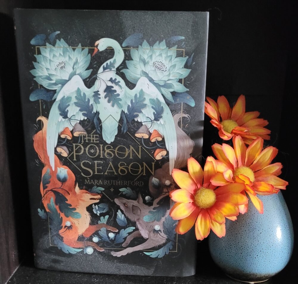 The Poison Season book, framed by flowers in a light blue vase.