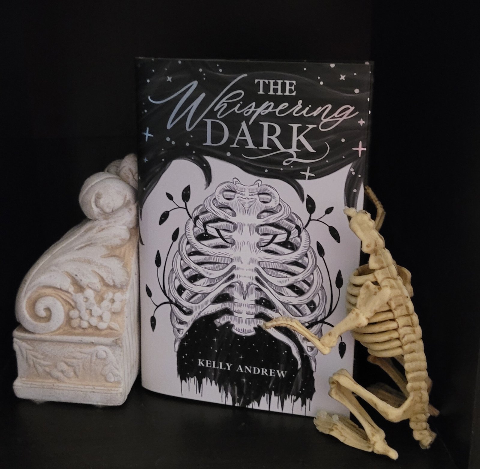 The Whispering Dark book framed by a skeleton and a floral relief carved bookend.